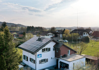 Haus mit Solarpanels im Vordergrund, dahinter weitere Häuser und Felder in Grün