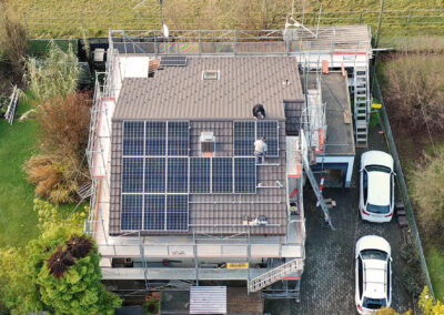 Zwei Männer montieren auf einem mit Gerüst eingepackten Haus mit Schrägdach Solarpanels. Aus Vogelperspektive.